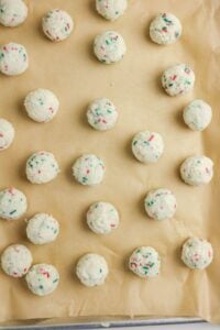 Unbaked cookie dough balls with colorful sprinkles are arranged in rows on a parchment-lined baking sheet.
