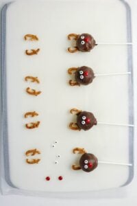 A baking sheet with three assembled chocolate reindeer cake pops and laid out pretzel antlers, candy eyes, and red candy noses.