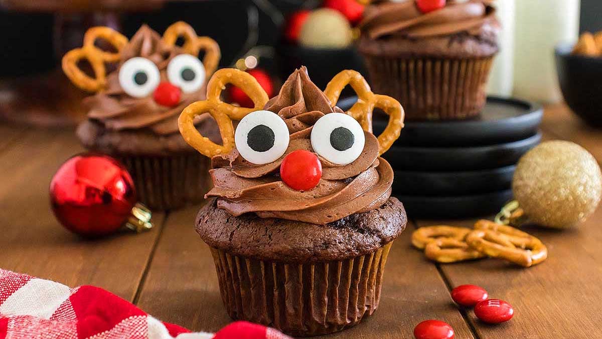 Chocolate cupcakes decorated with chocolate frosting, candy eyes, red candy noses, and pretzel antlers, resembling reindeer, are arranged on a wooden table with festive ornaments.