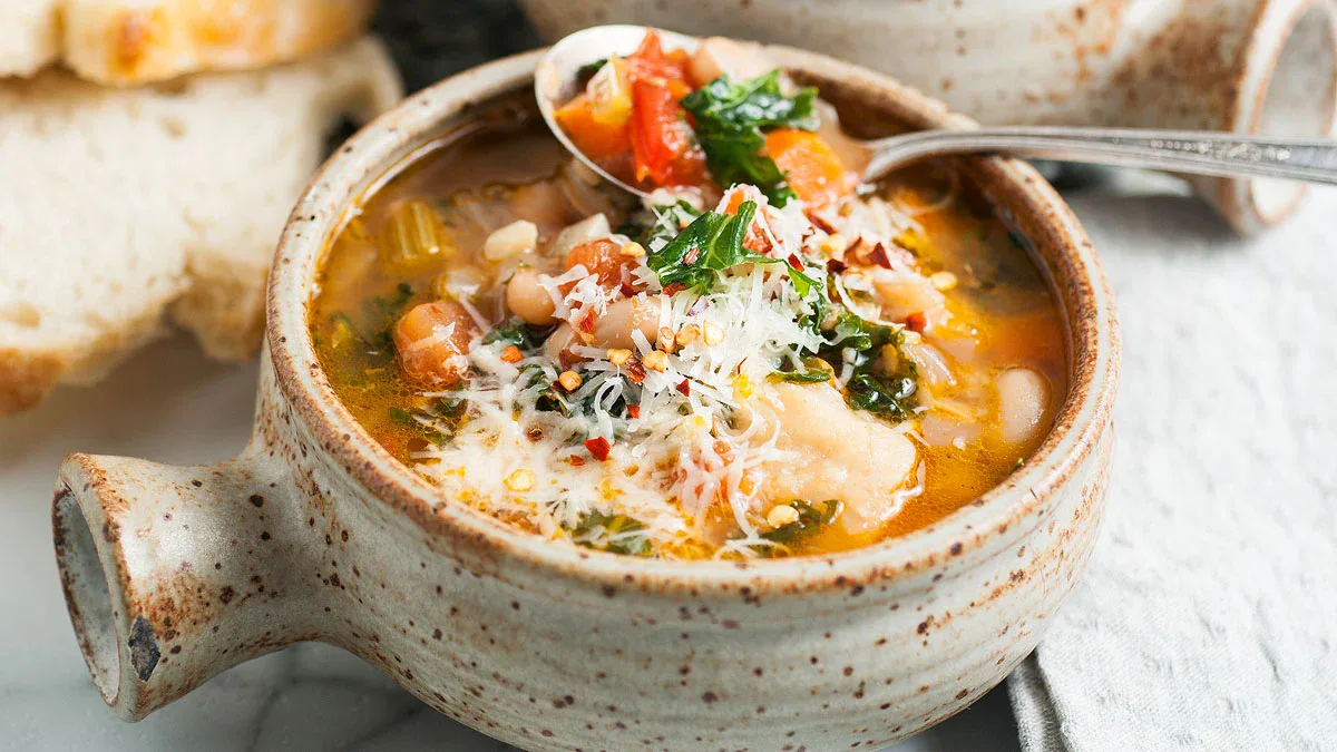 A rustic ceramic bowl filled with vegetable and bean soup, topped with grated cheese and herbs, with a spoon and slices of bread beside it.