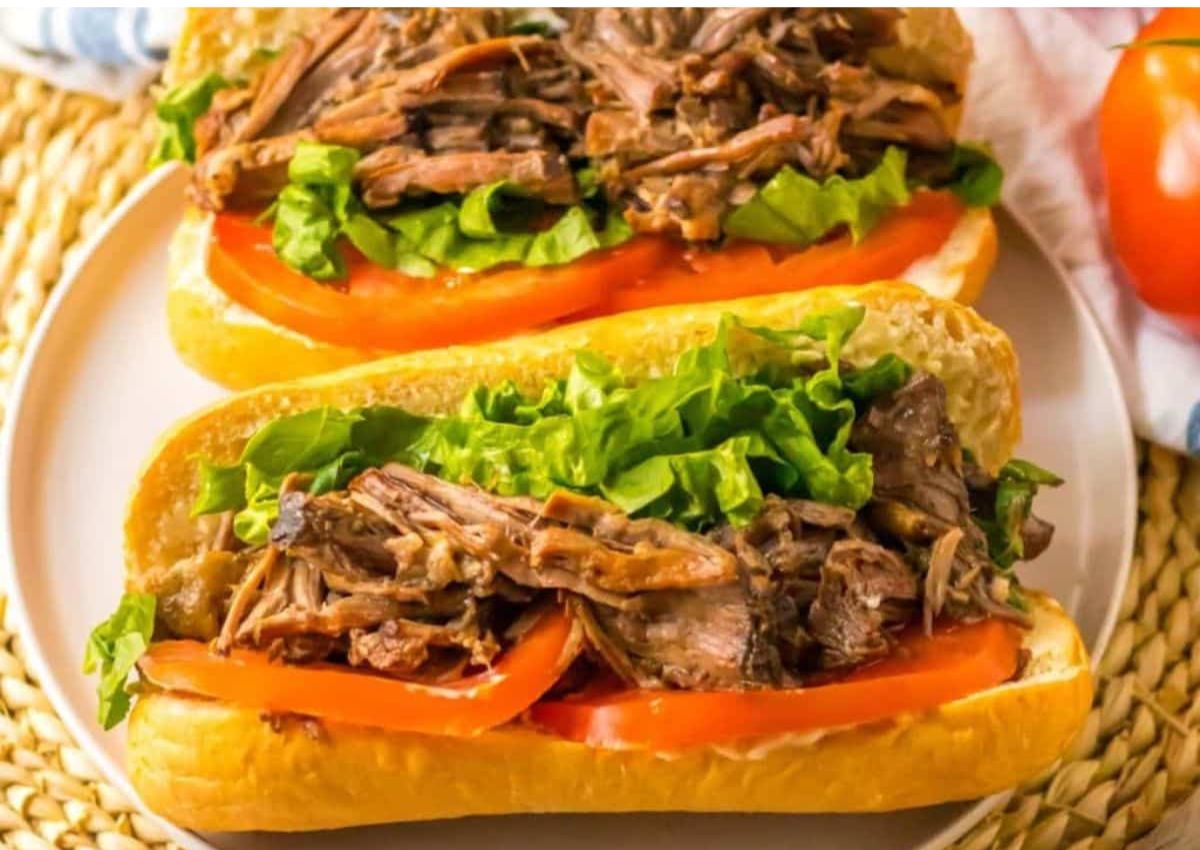 Two sandwiches on a plate filled with shredded meat, lettuce, and tomato slices in hoagie rolls, placed on a woven placemat.