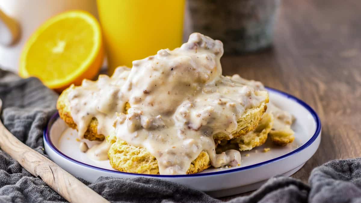 A plate of biscuits topped with sausage gravy sits on a table, with a halved orange and a glass of juice in the background.