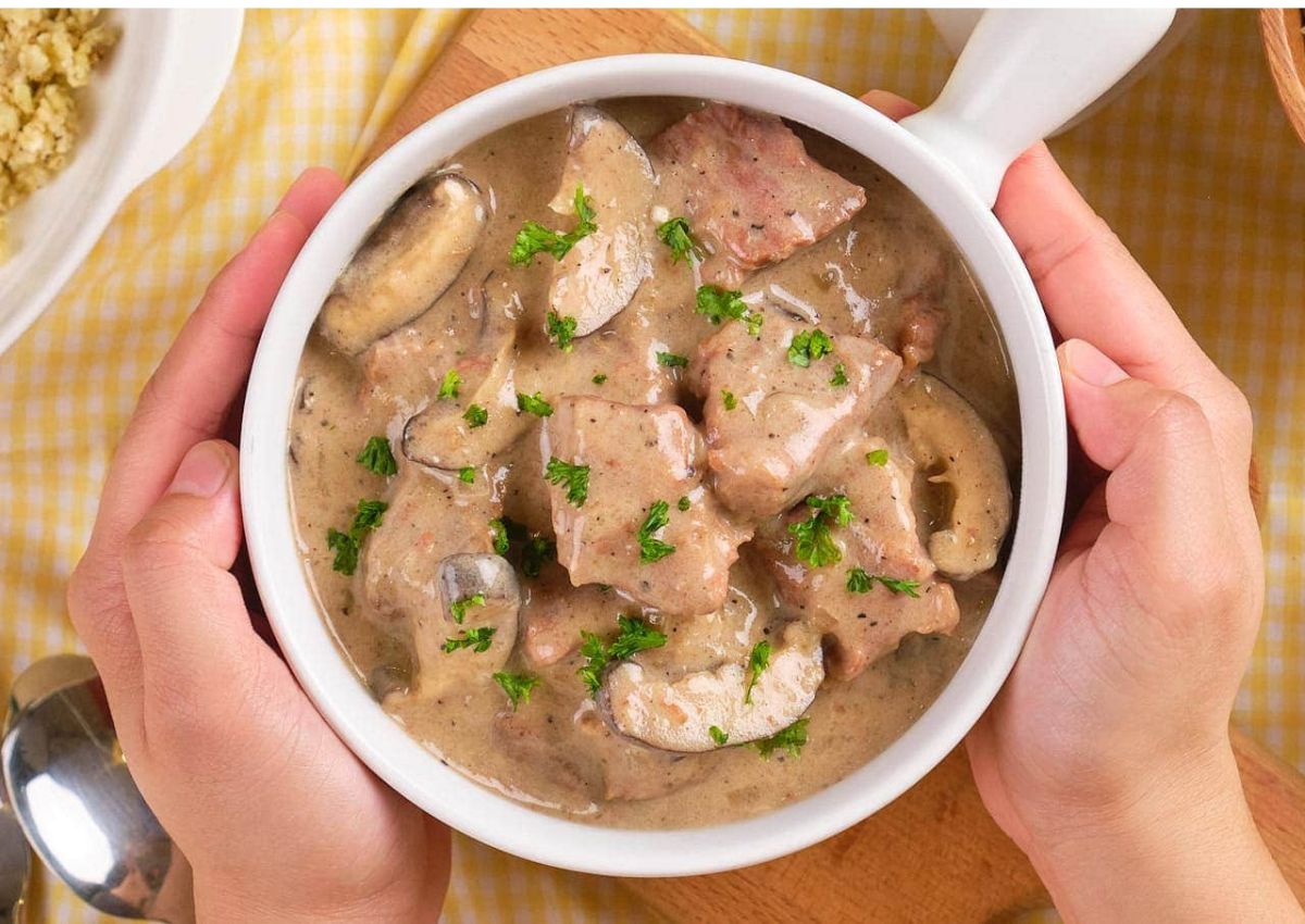 A bowl of beef stroganoff with mushrooms in a creamy sauce, garnished with chopped parsley, held by two hands.