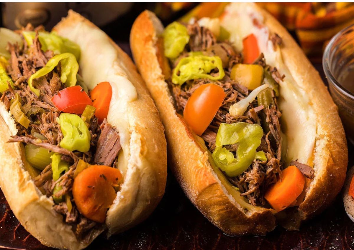 Two hoagie rolls filled with shredded beef, sliced peppers, carrots, and melted cheese, served with a side of dipping sauce.