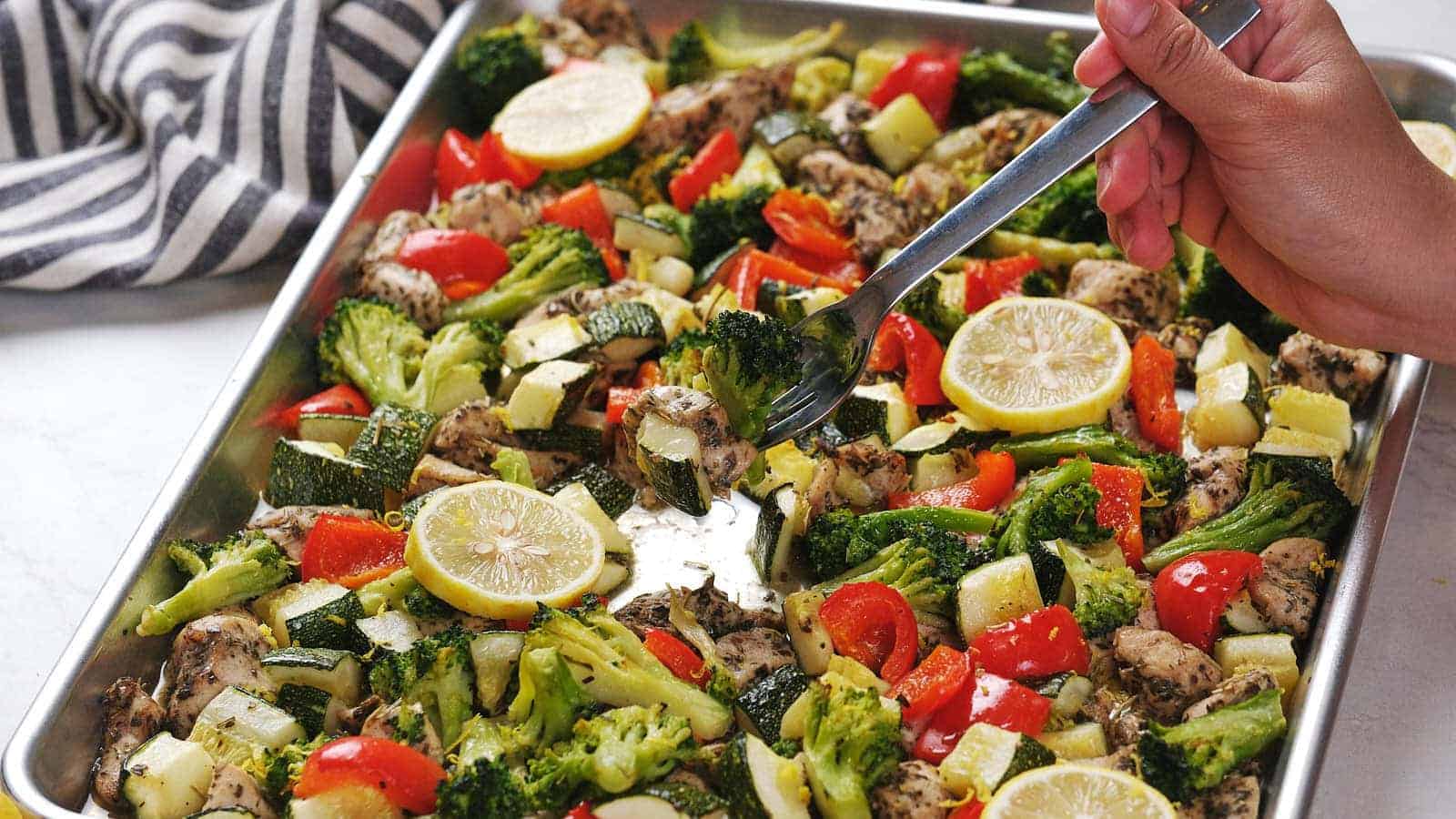 A hand holds a spoon over a baking sheet filled with roasted vegetables, chicken pieces, and lemon slices.