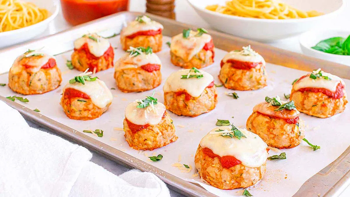 A baking sheet with chicken parmesan meatballs topped with marinara sauce, melted cheese, and chopped herbs on parchment paper. Spaghetti and sauce are visible in the background.