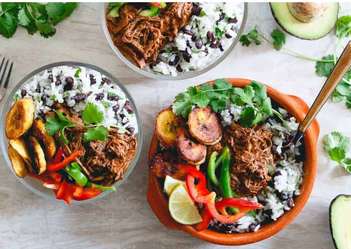 Three bowls filled with rice, black beans, shredded meat, fried plantains, sliced peppers, cilantro, and lime wedges, with avocado and cilantro garnishes nearby.