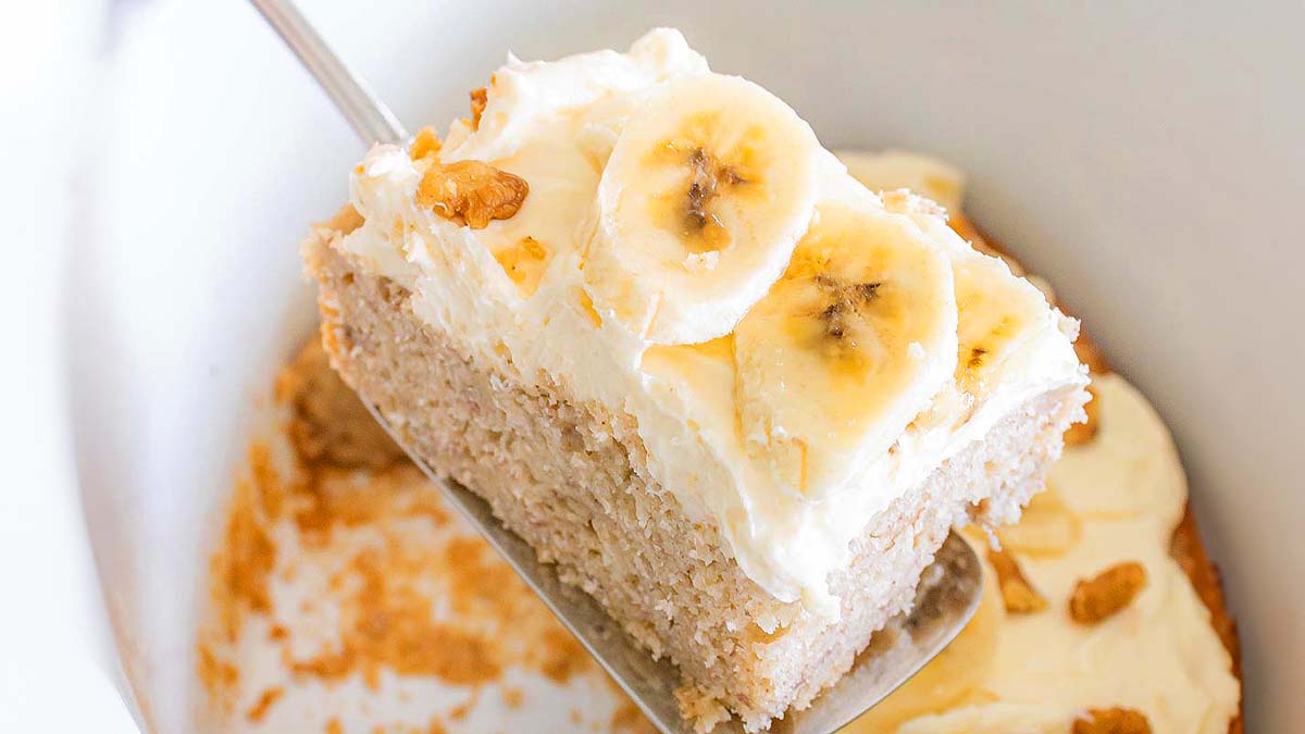 A slice of banana cake with cream cheese frosting and banana slices on top, being lifted from a baking dish with a spatula.