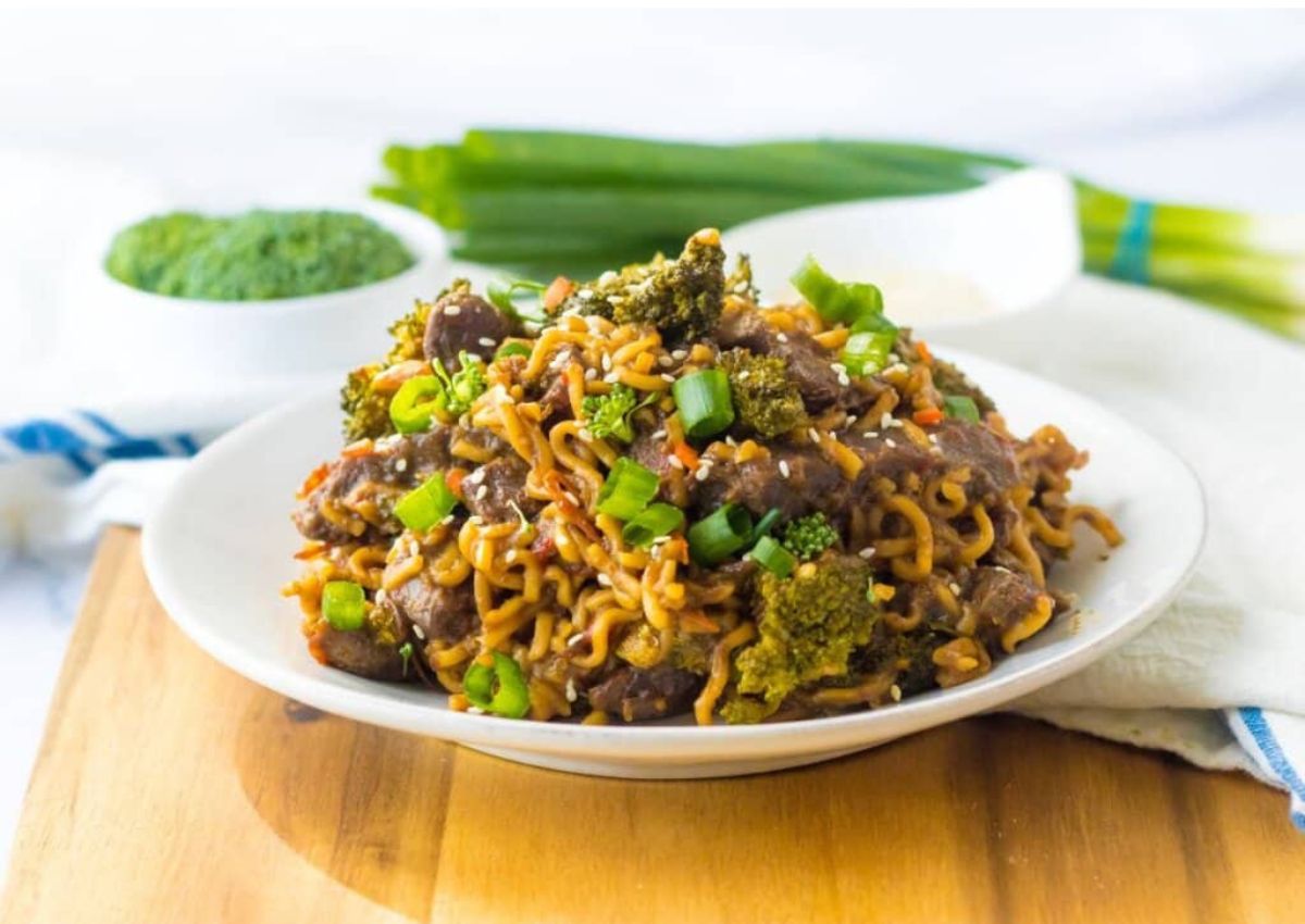 A plate of stir-fried noodles with beef, broccoli, and green onions, garnished with sesame seeds, sits on a wooden board with fresh green onions in the background.