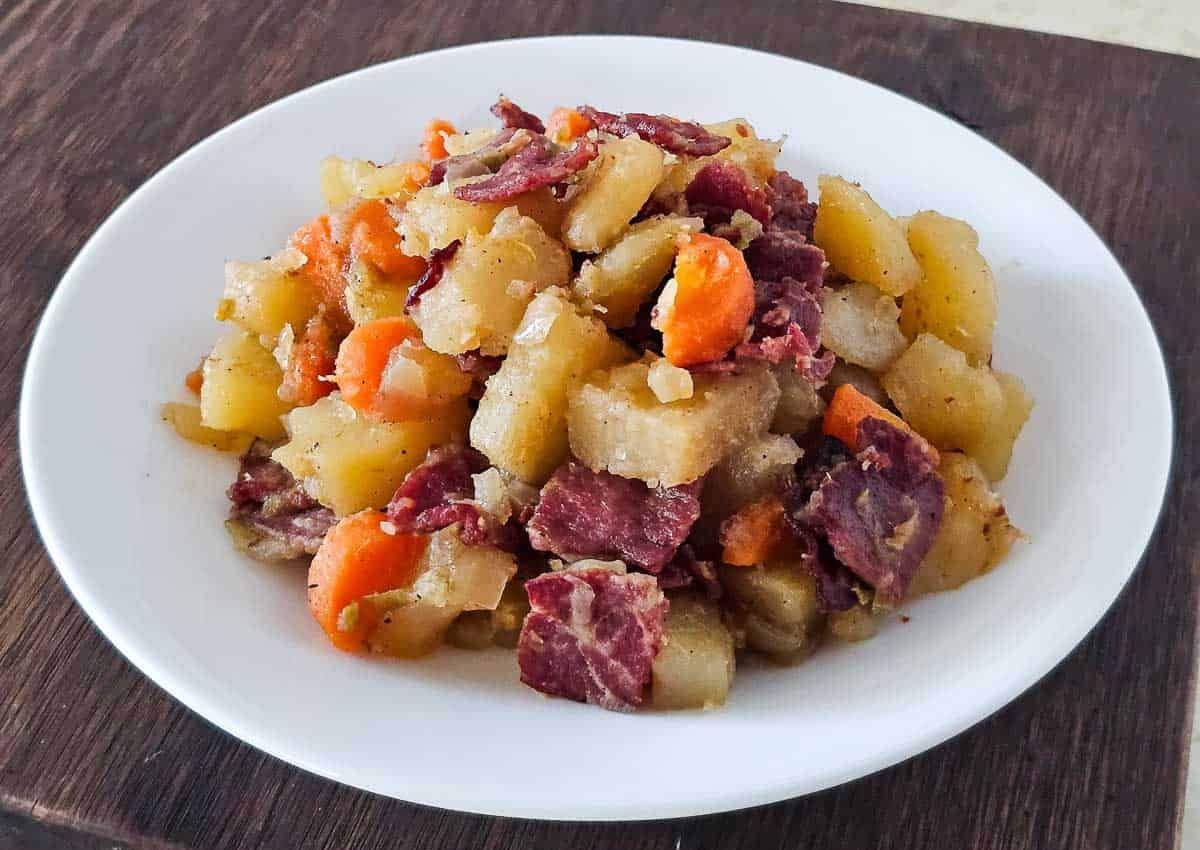 A white plate with a serving of corned beef hash containing diced potatoes, carrot slices, and pieces of corned beef on a wooden surface.