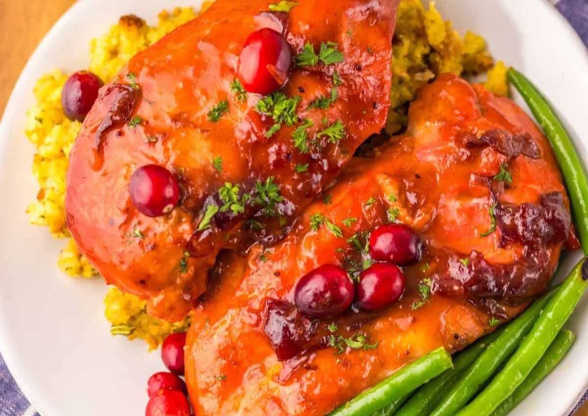 Two glazed chicken breasts topped with cranberries and herbs, served on yellow rice with a side of green beans on a white plate.