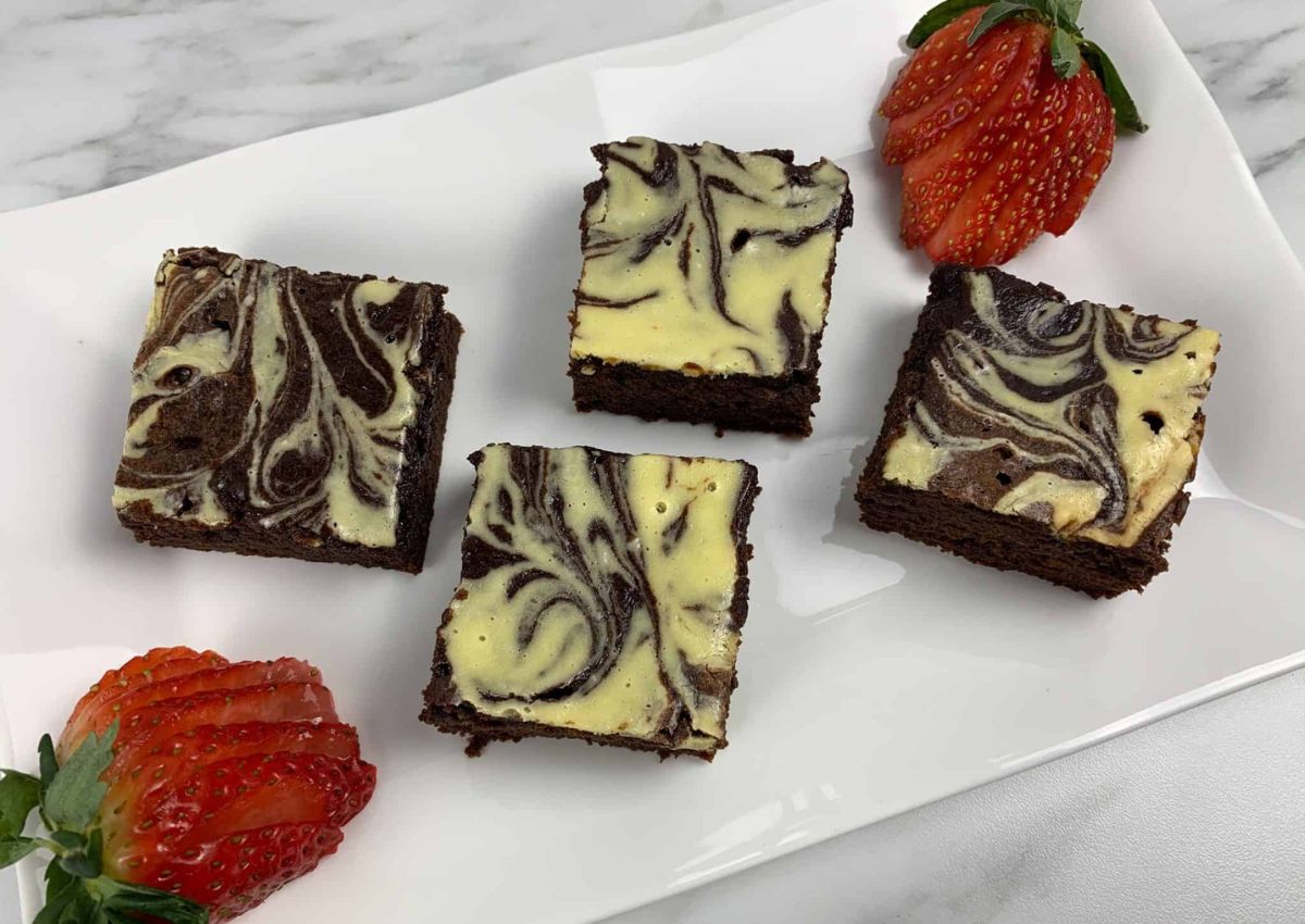 Four cheesecake brownies with a marbled pattern are arranged on a white plate, accompanied by three whole and halved strawberries.