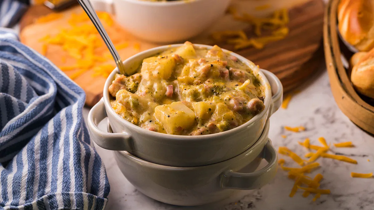 A bowl of creamy potato and vegetable soup with a spoon, surrounded by shredded cheese, a striped napkin, and bread rolls.