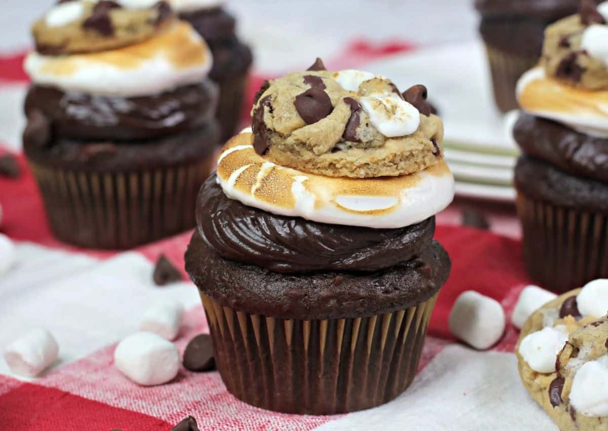 Chocolate cupcakes topped with chocolate frosting, toasted marshmallow, and a chocolate chip cookie, with mini marshmallows and chocolate chips scattered nearby on a checkered tablecloth.