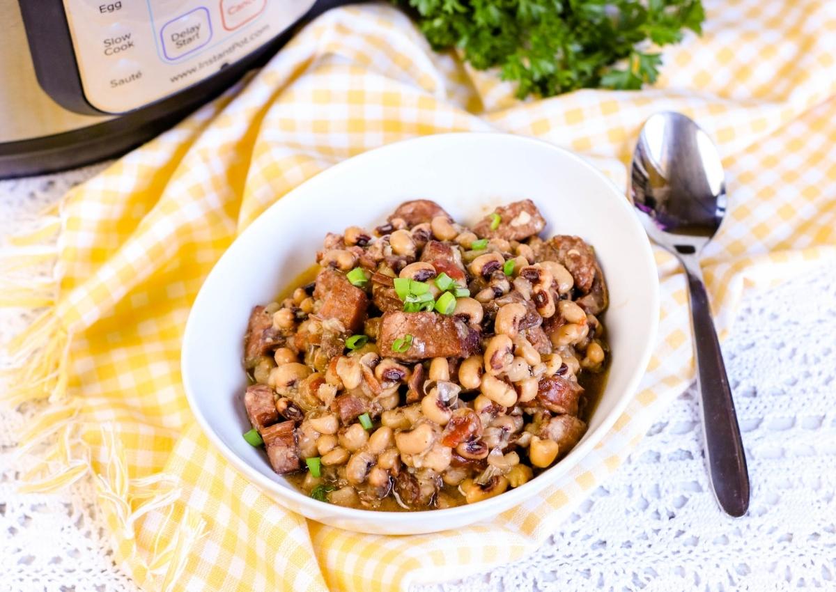 A white bowl filled with cooked black-eyed peas and chunks of meat, garnished with chopped green onions, sits on a yellow checkered cloth next to a spoon and an Instant Pot.