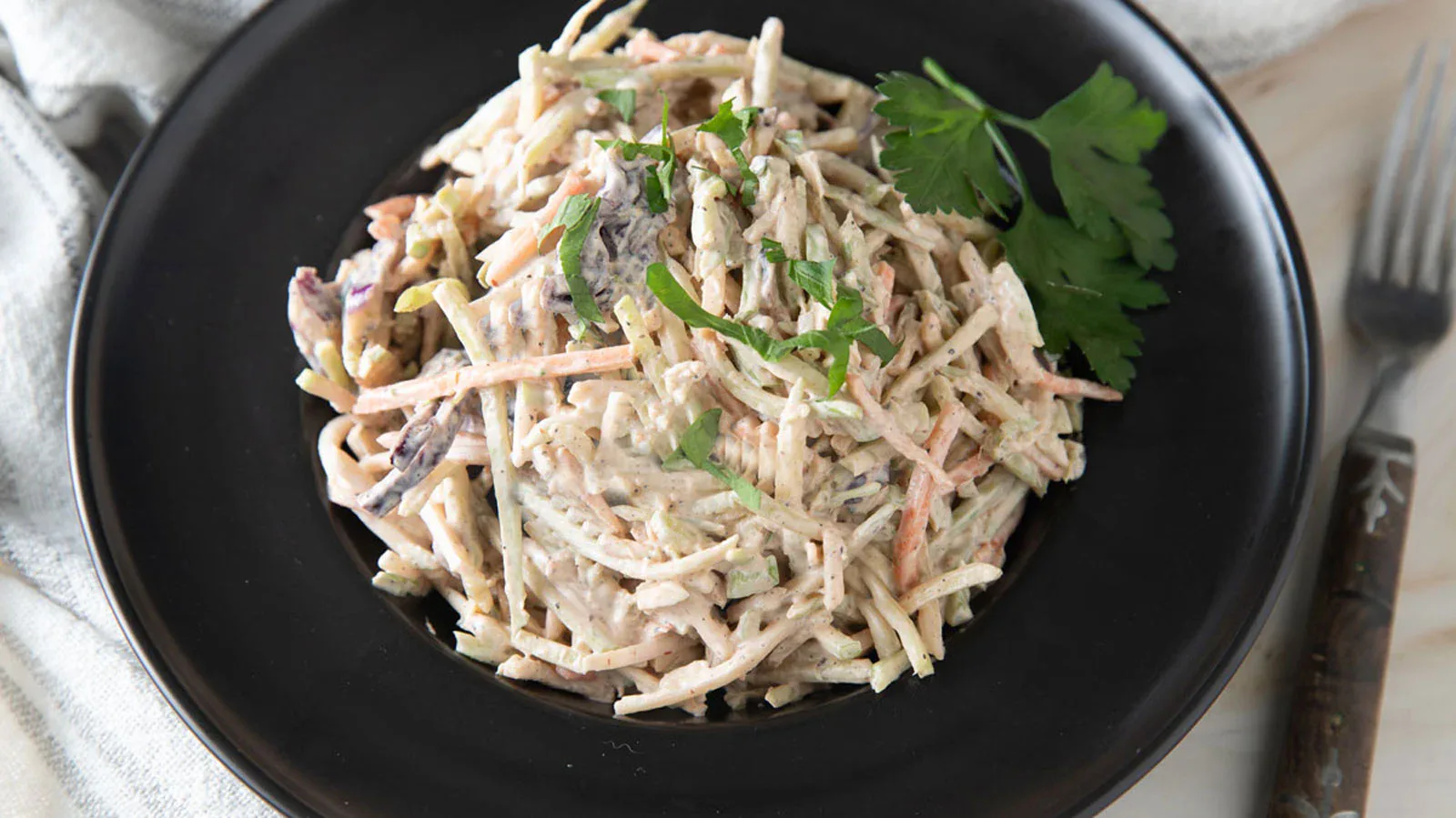 A black plate with a serving of creamy coleslaw garnished with fresh parsley, next to a fork on a white surface.