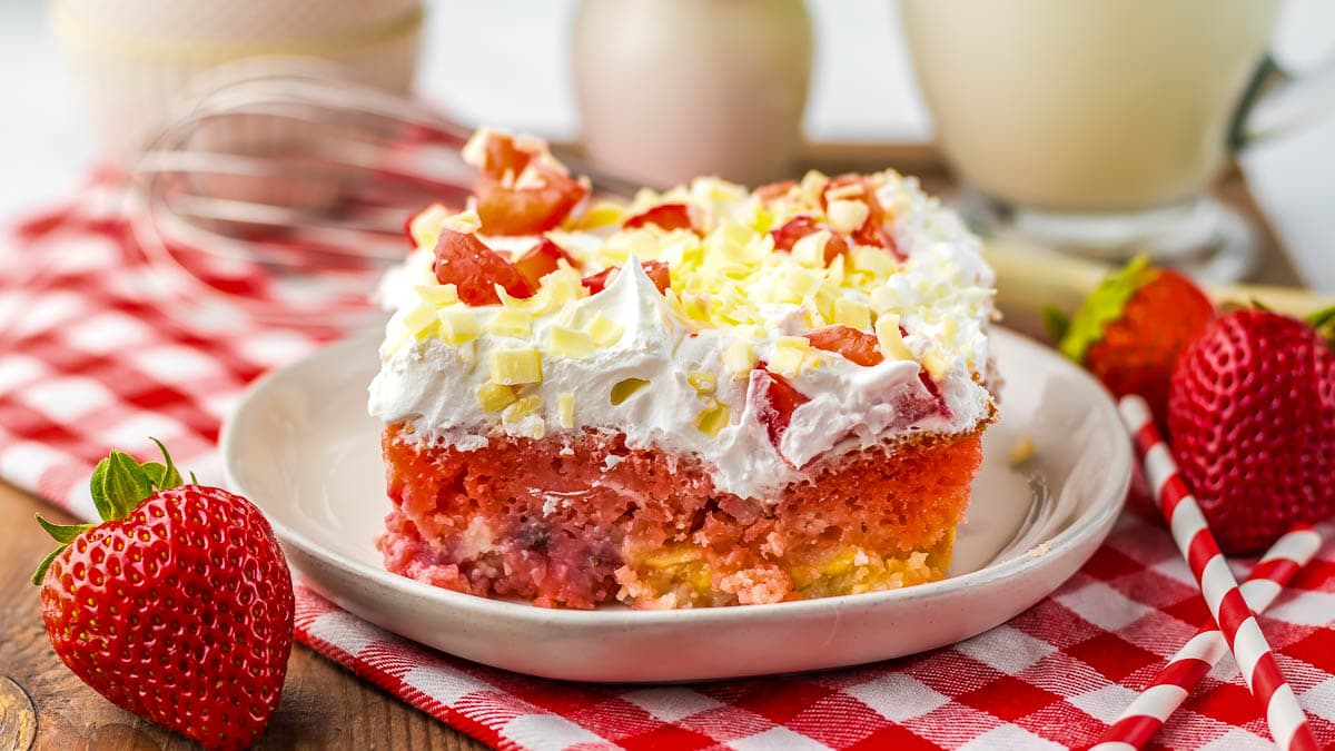 A slice of strawberry and cream cake topped with whipped cream and white chocolate, served on a plate with fresh strawberries, on a red checkered cloth.
