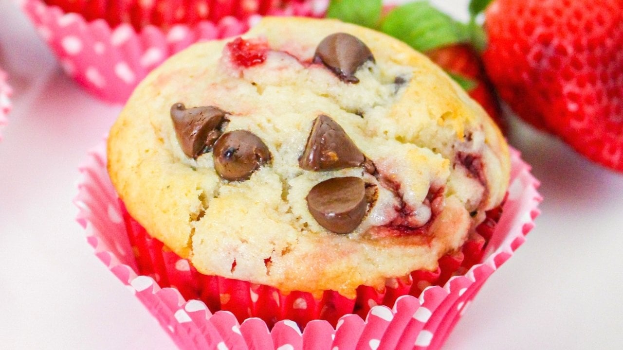 A muffin with chocolate chips and visible pieces of strawberry in a pink polka dot wrapper, placed next to fresh strawberries.