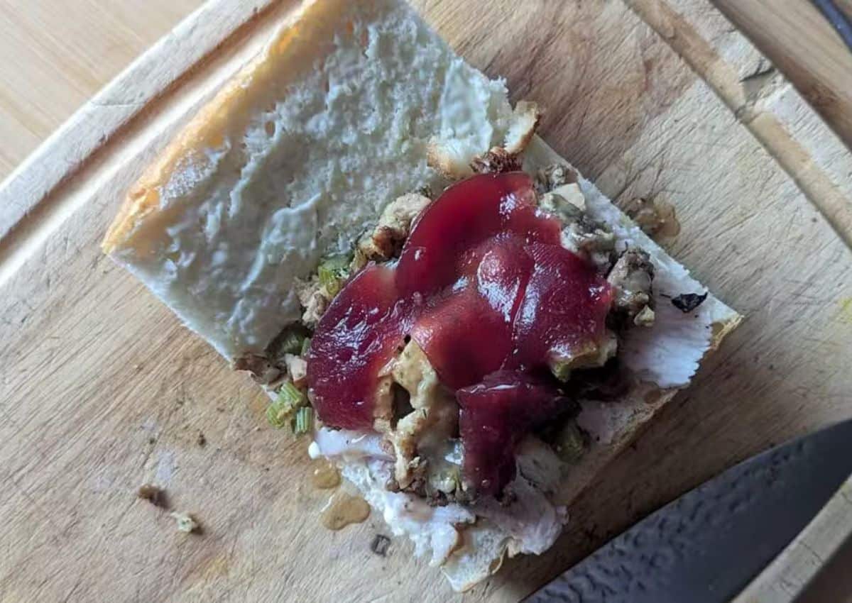 A slice of bread topped with chopped stuffing, cranberry sauce, and a spread, sitting on a wooden cutting board next to a knife.