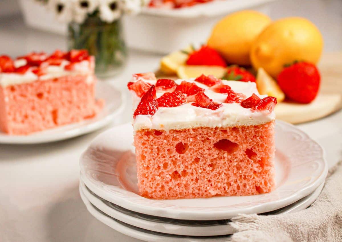 A slice of strawberry cake with whipped frosting and diced strawberries on top, served on a white plate, with lemons and whole strawberries in the background.
