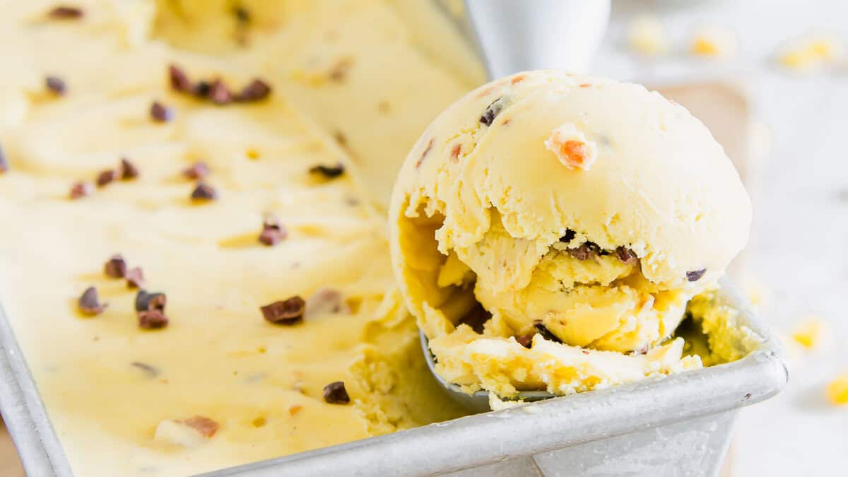 A close-up of a metal pan filled with creamy yellow ice cream, with a scoop resting on the edge and small chunks mixed throughout.