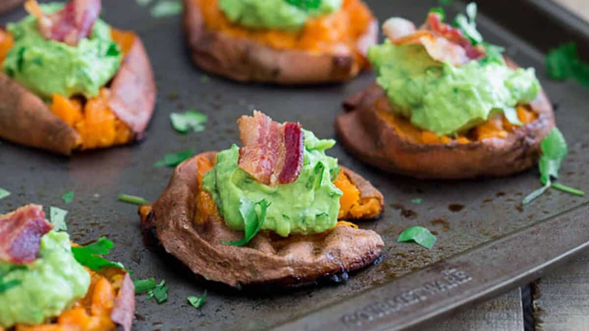 Baked sweet potato rounds topped with guacamole, chopped cilantro, and pieces of bacon on a baking sheet.