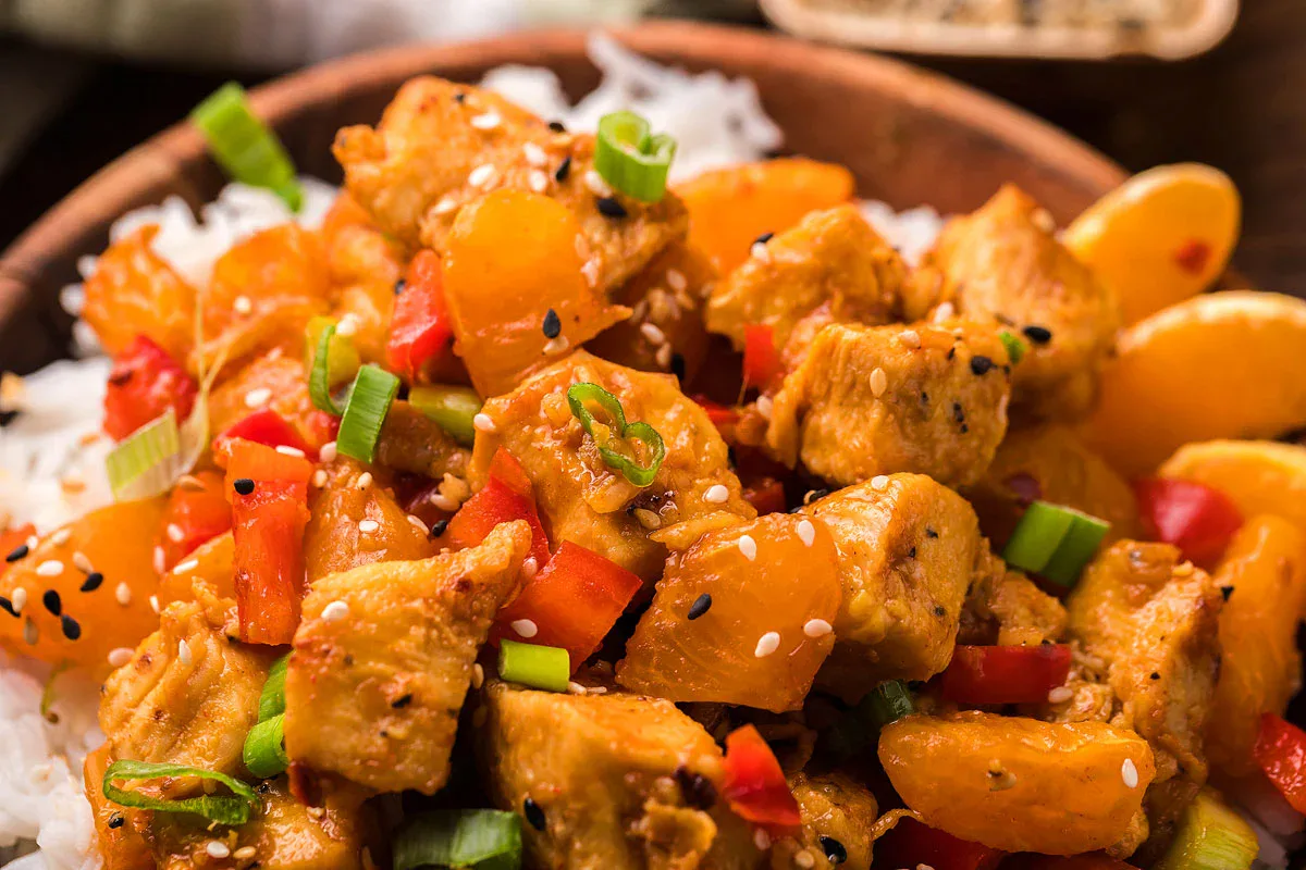 A bowl of white rice topped with pieces of cooked chicken, orange segments, diced red bell peppers, sliced green onions, and sprinkled with sesame seeds.