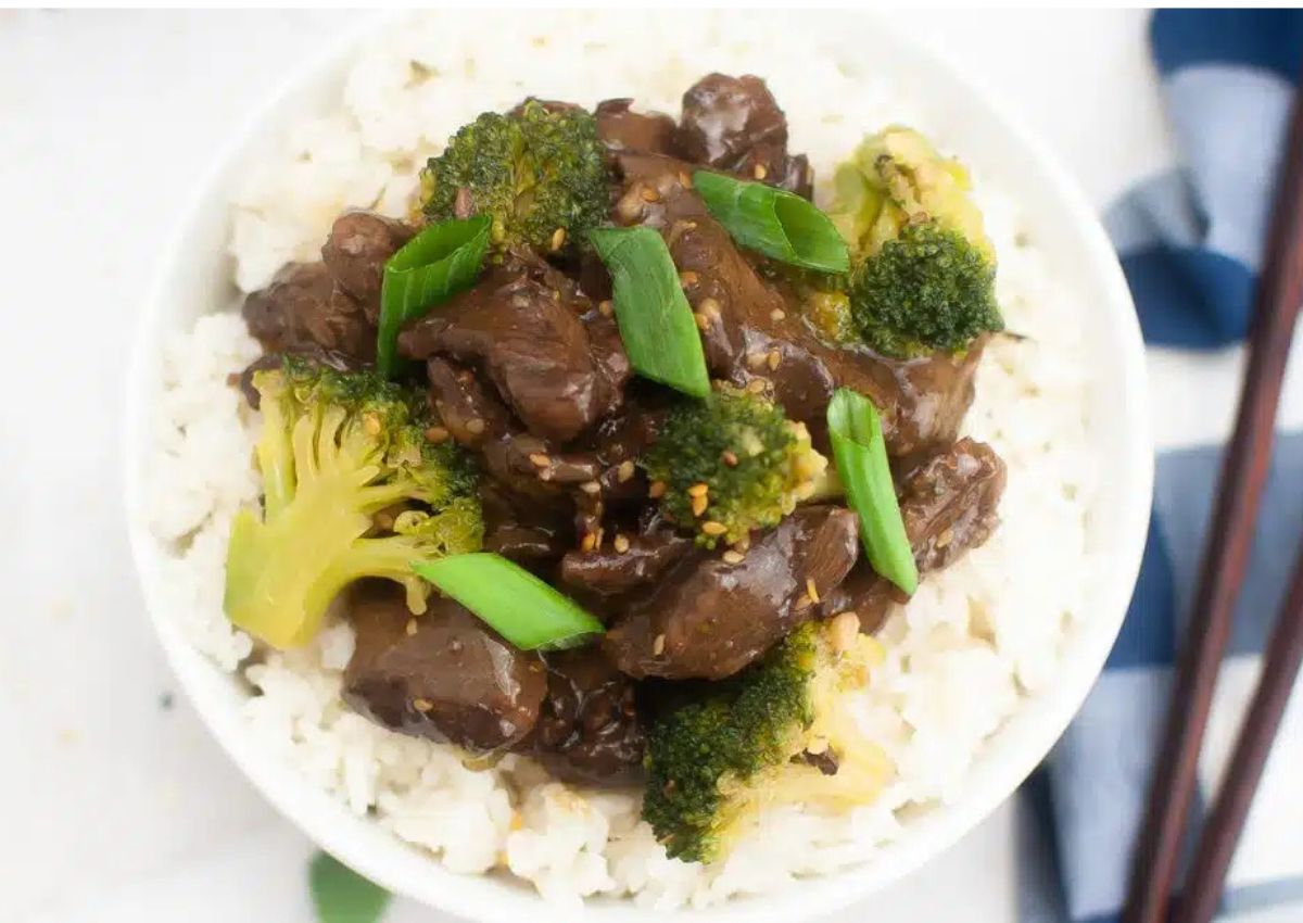 A bowl of white rice topped with beef, broccoli, and sliced green onions, served with chopsticks on the side.