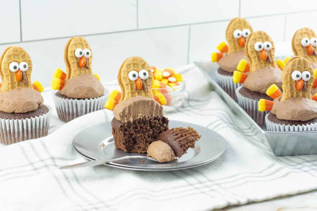 Chocolate cupcakes decorated as turkeys with cookie and candy toppings, one cut open on a plate with a fork, others arranged in a tray in the background.