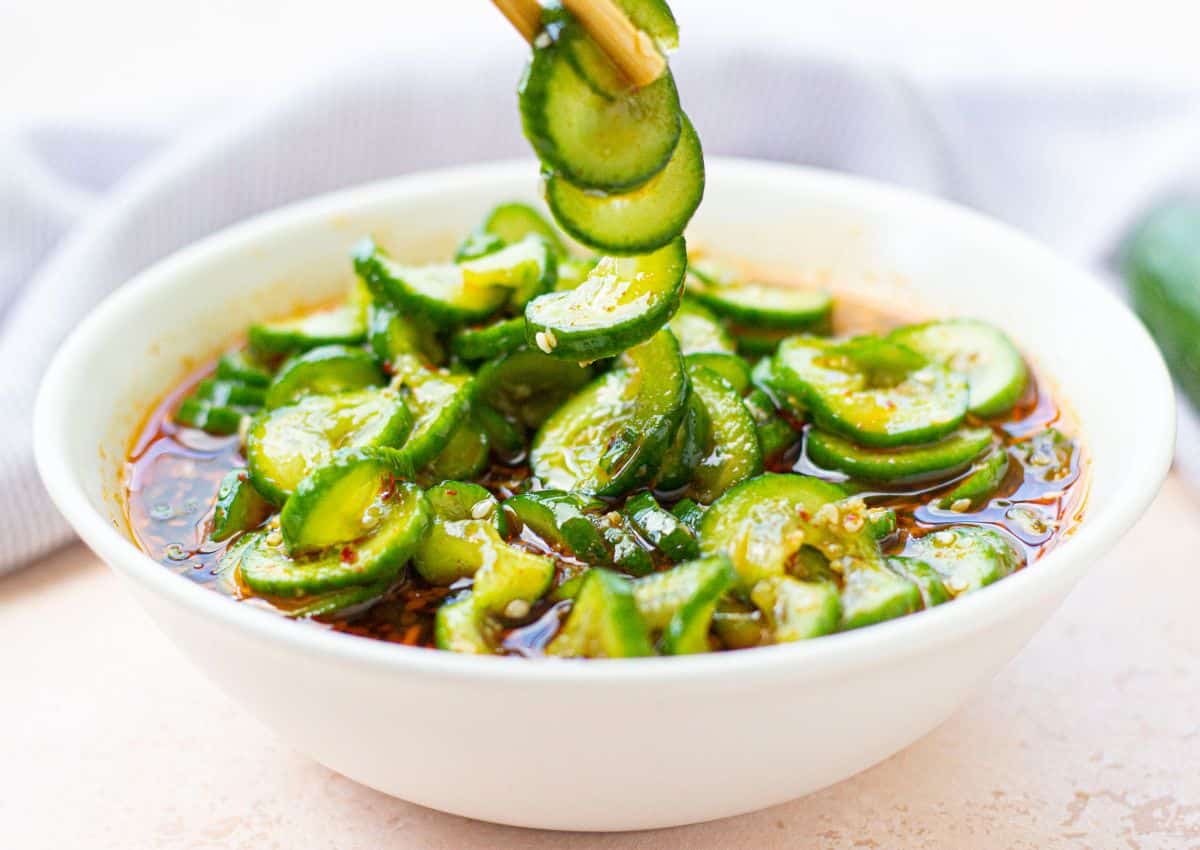 A white bowl filled with sliced cucumbers in a chili oil sauce, with chopsticks lifting some cucumber slices.