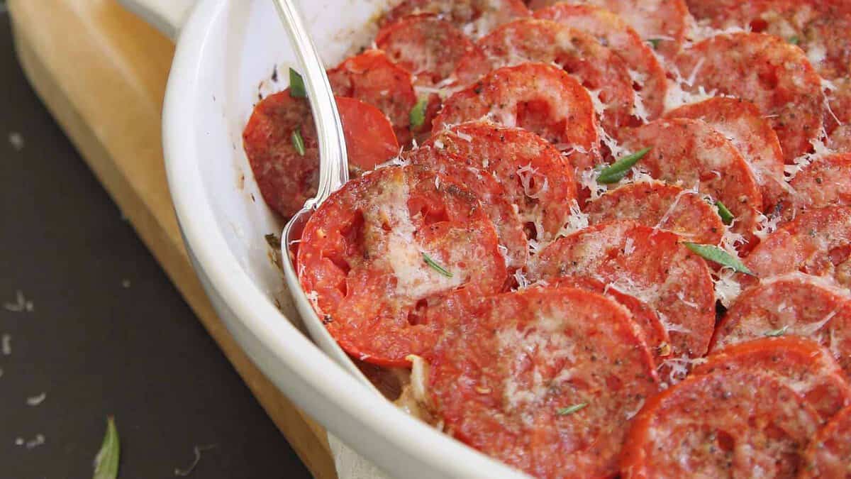 A close-up of a baked tomato casserole topped with grated cheese, herbs, and black pepper in a white baking dish with a serving spoon.