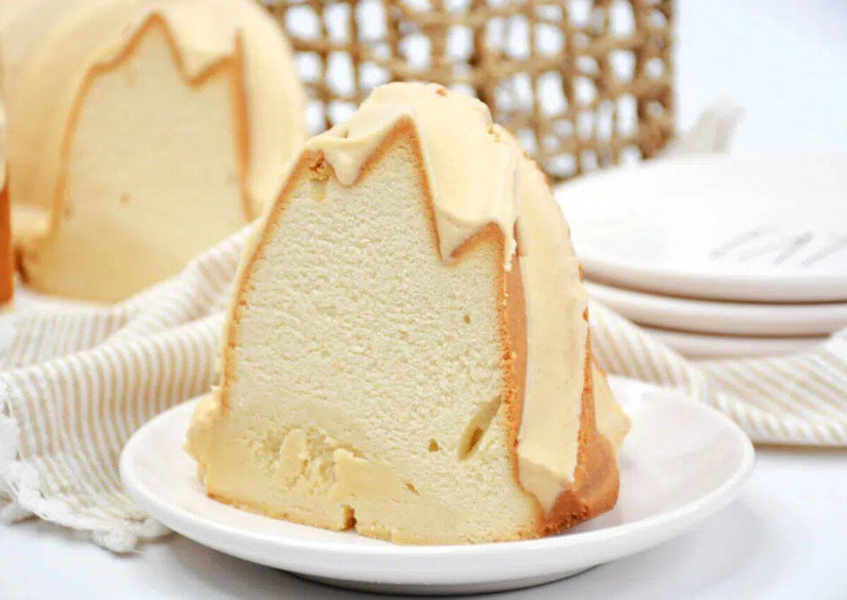 A thick slice of plain pound cake with a light glaze sits on a white plate, with a bundle of plates and a striped cloth napkin in the background.