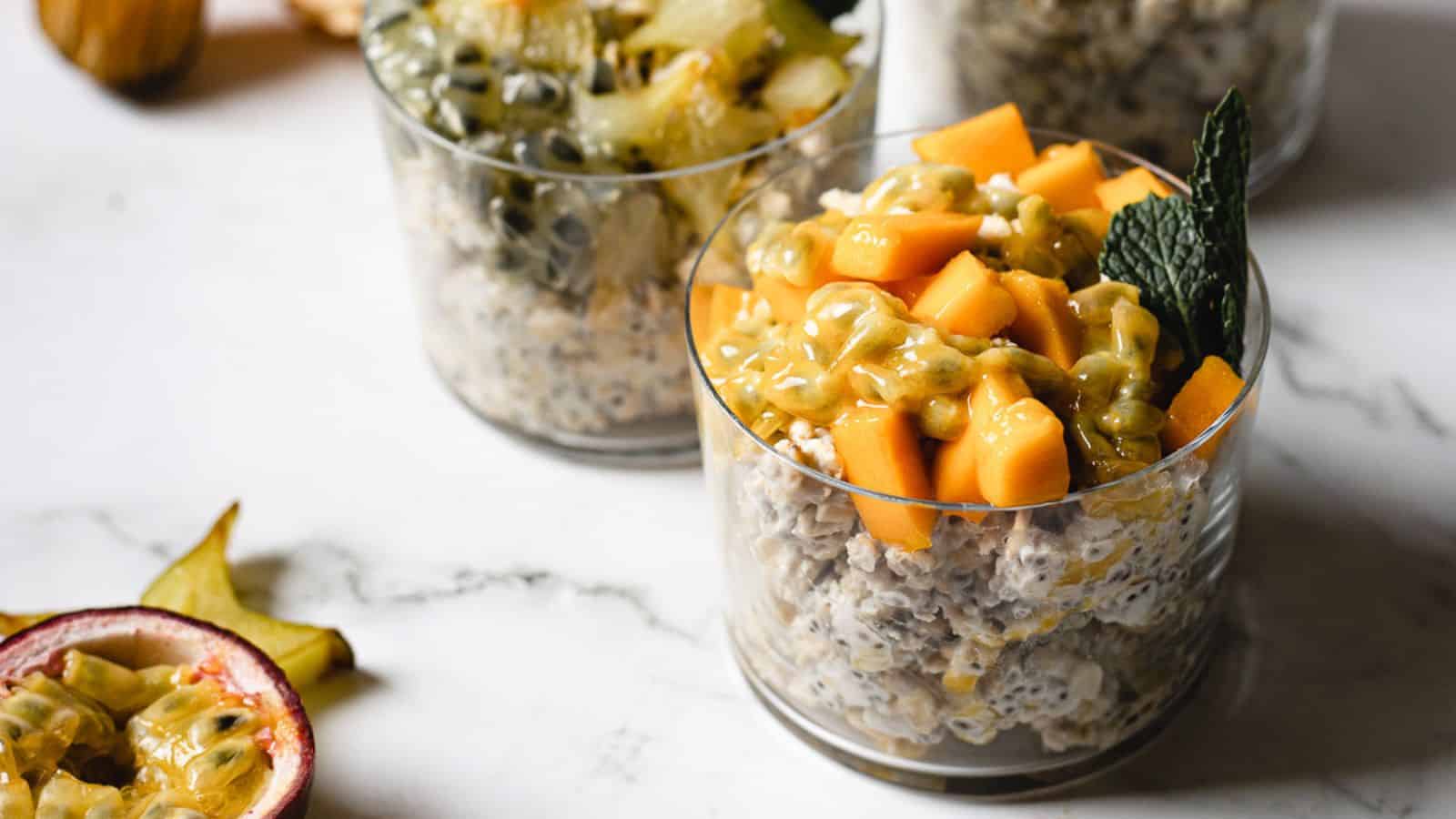Three glasses filled with chia pudding, topped with chopped fresh fruits including mango, passion fruit, and starfruit, placed on a marble surface.