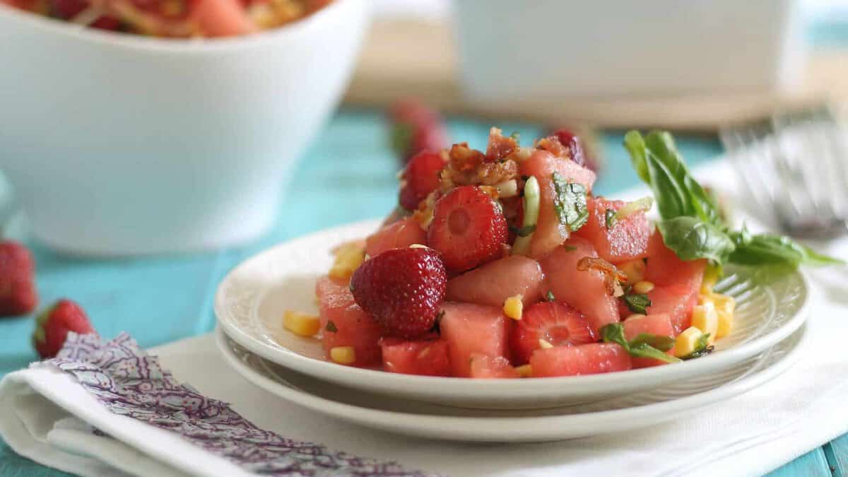 A plate of watermelon and strawberry salad garnished with basil and corn, placed on a white napkin with a fork in the background.