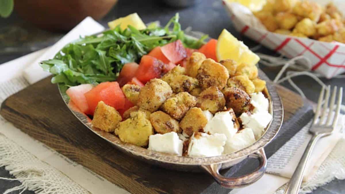 A bowl containing diced watermelon, arugula, lemon wedges, fried cauliflower, and cubes of feta cheese, placed on a wooden board with a fork beside it.