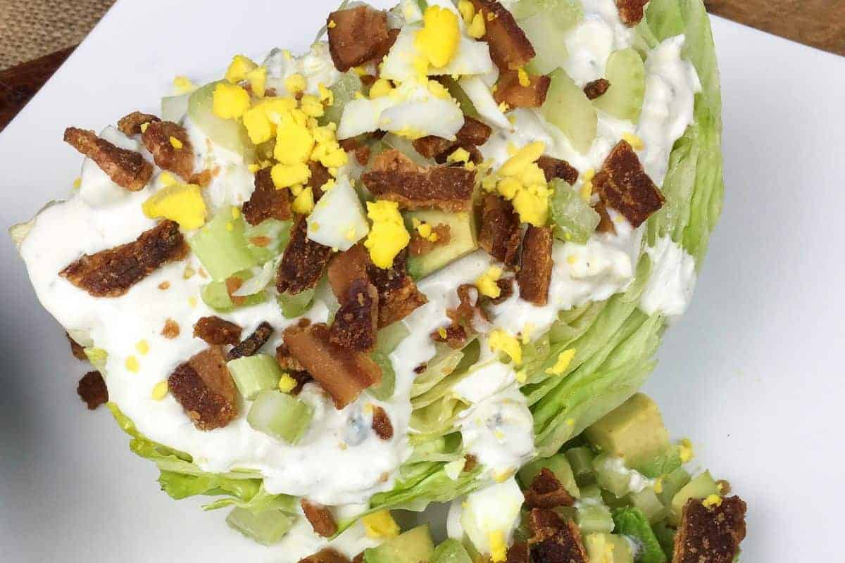 A wedge of iceberg lettuce topped with chopped hard-boiled egg, bacon pieces, diced celery, and creamy dressing on a white plate.