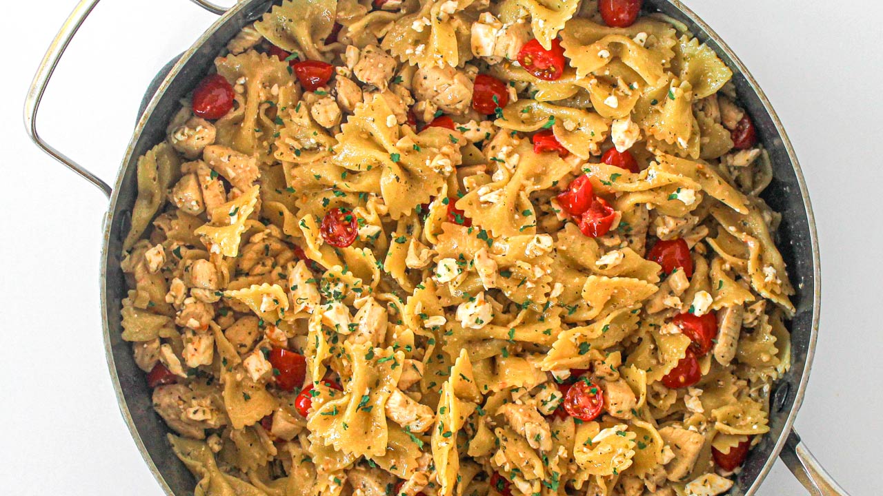 A pan filled with bowtie pasta, diced chicken, cherry tomatoes, crumbled cheese, and herbs.