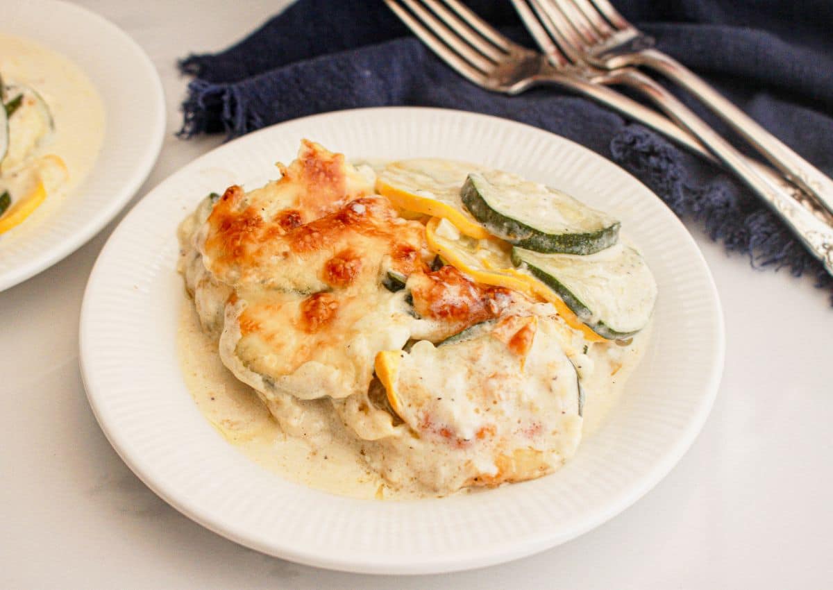 A plate of creamy baked squash and zucchini gratin topped with melted cheese, served on a white dish with a navy napkin and metal forks in the background.