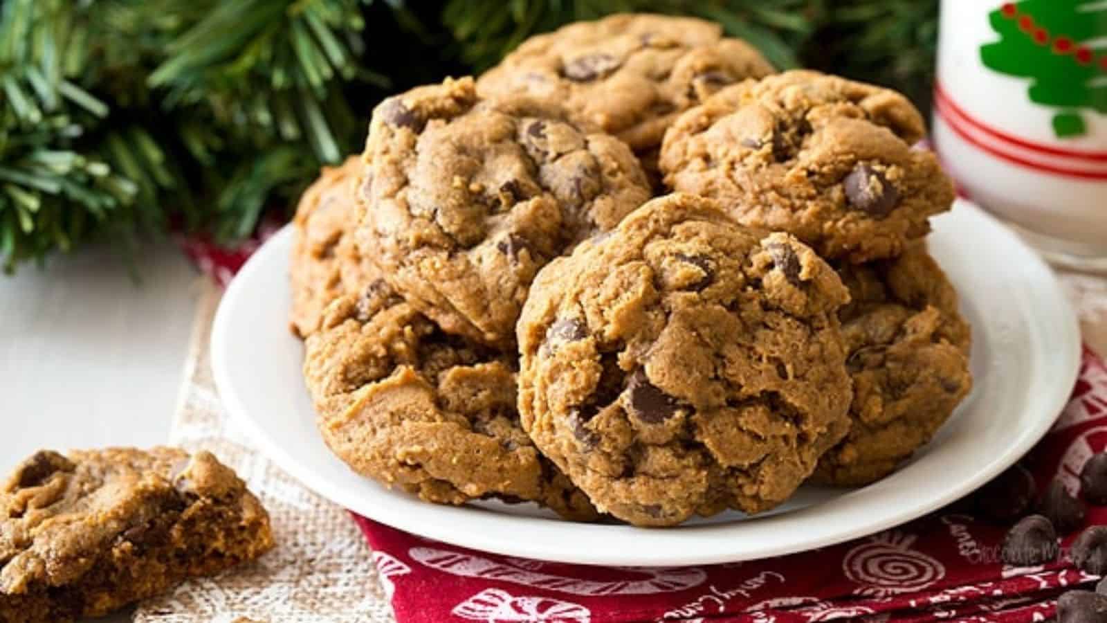 A white plate with several chocolate chip cookies sits on a festive red napkin near a glass of milk and holiday greenery.