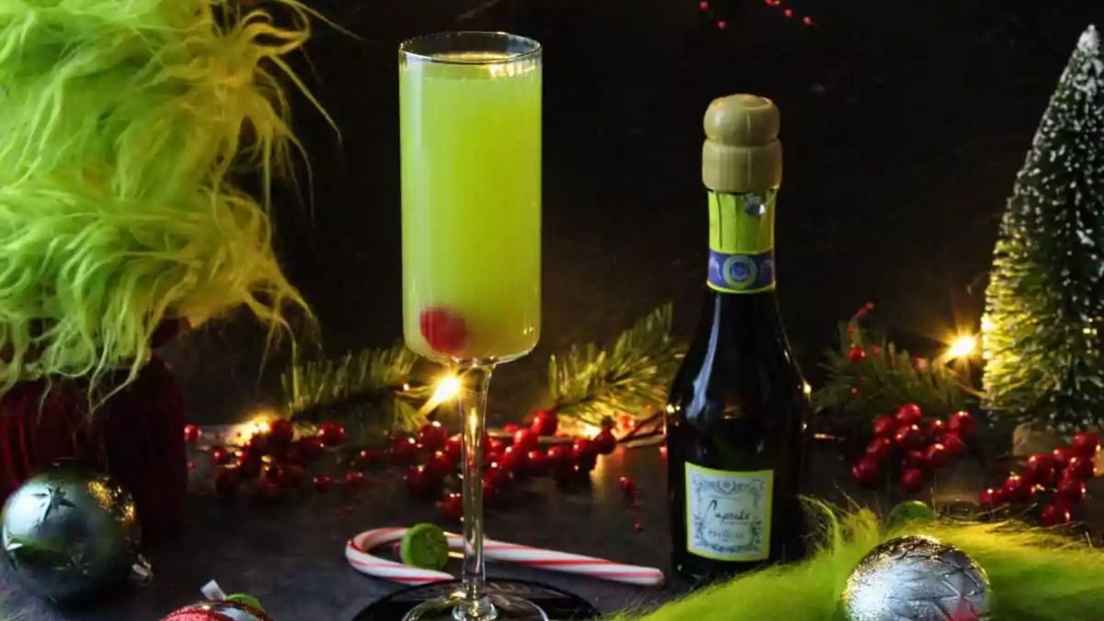 A tall champagne glass with a yellow-green drink and a red cherry, next to a mini champagne bottle, surrounded by festive holiday decorations and green faux fur.