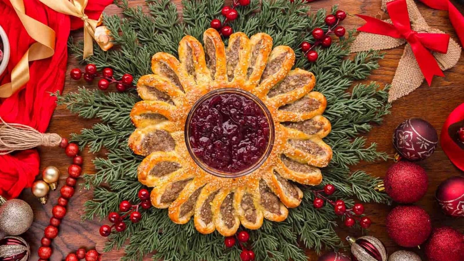 A festive pastry shaped like a sunflower with a bowl of cranberry sauce in the center, surrounded by evergreen branches and Christmas ornaments.