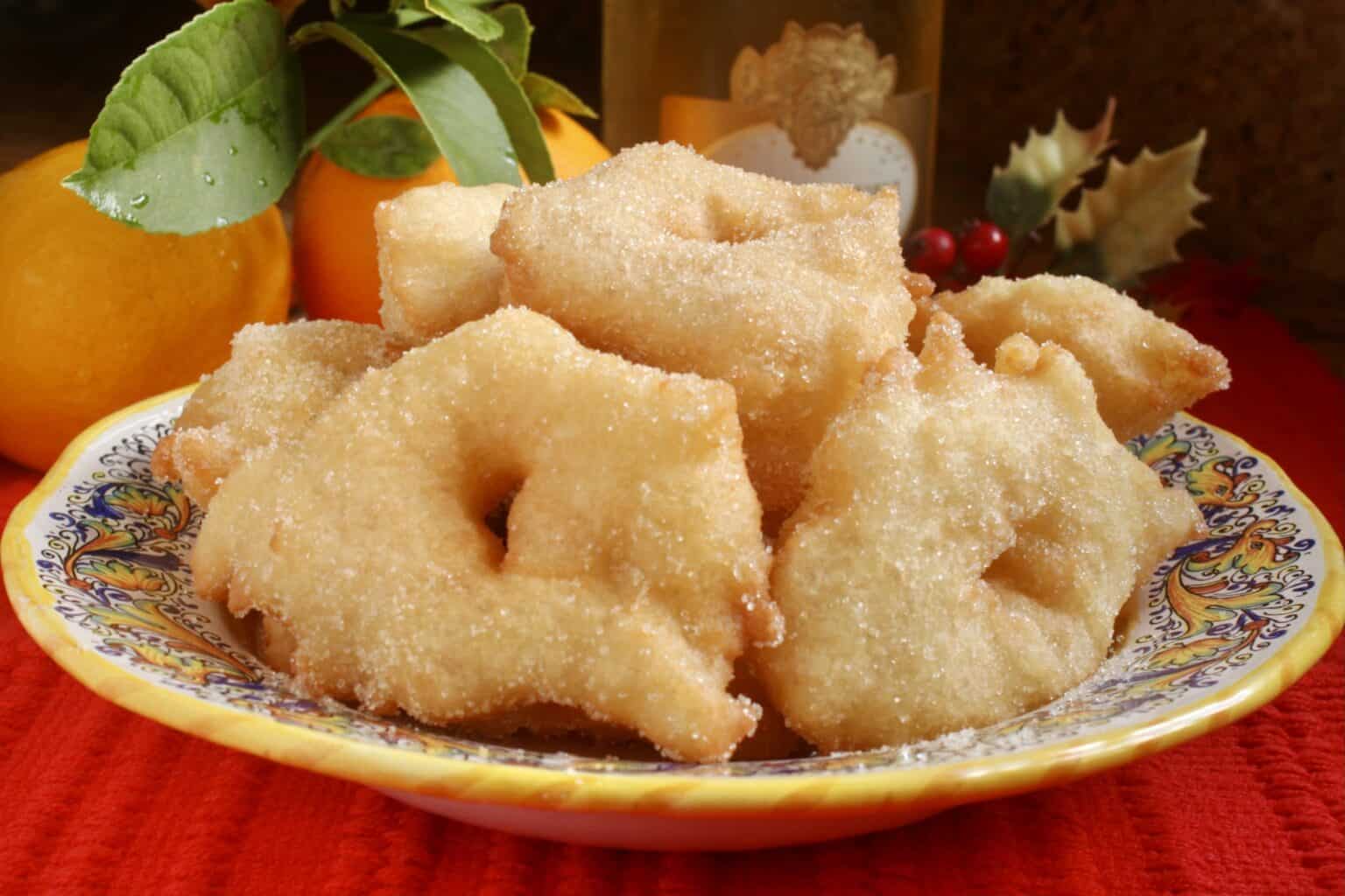 A plate of sugar-coated, ring-shaped fritters sits on a patterned dish, with oranges and green leaves in the background.
