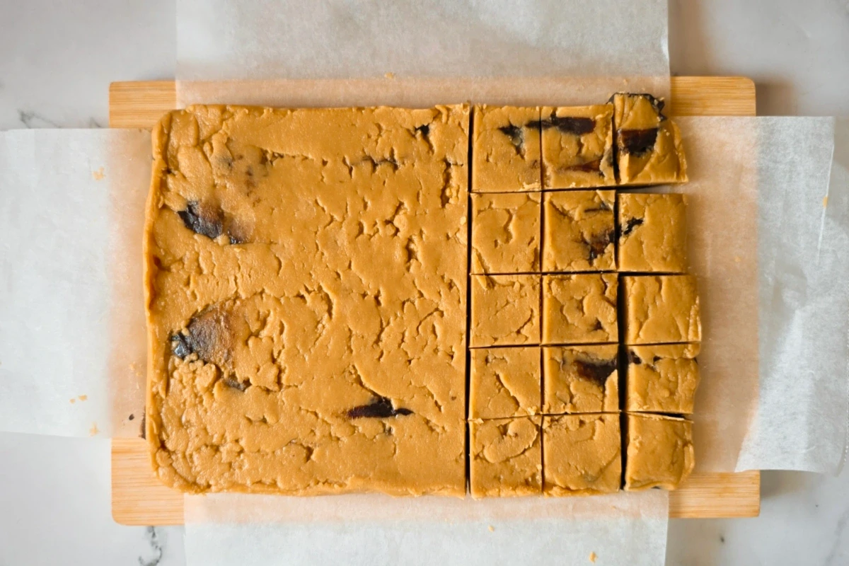 Rectangular slab of cookie dough with chocolate chunks, partially cut into squares, sitting on parchment paper atop a wooden board.