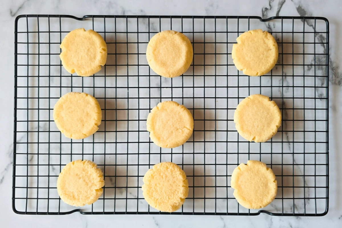Nine round, yellow cookies are evenly spaced on a black wire cooling rack set on a white marble surface.