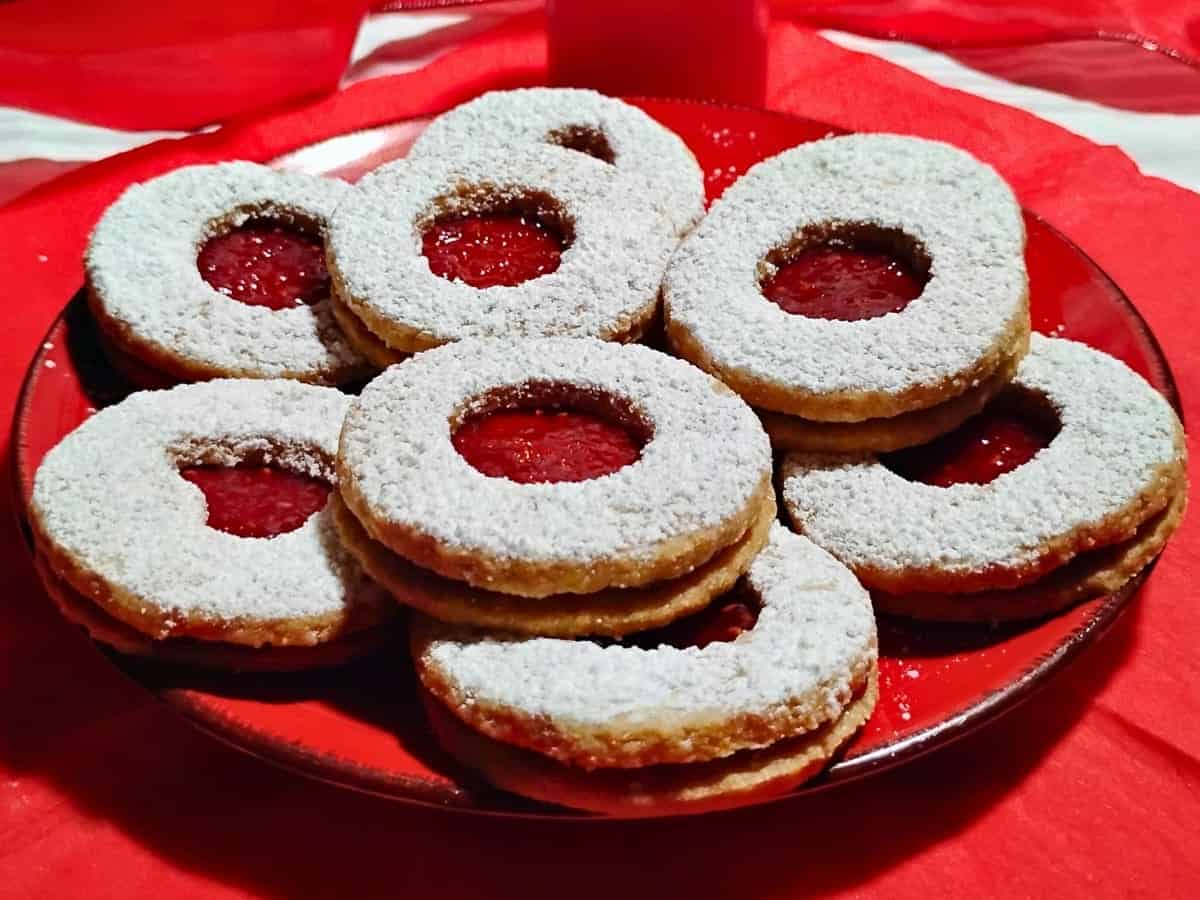 A plate of round Linzer cookies filled with red jam and dusted with powdered sugar, arranged on a red plate.
