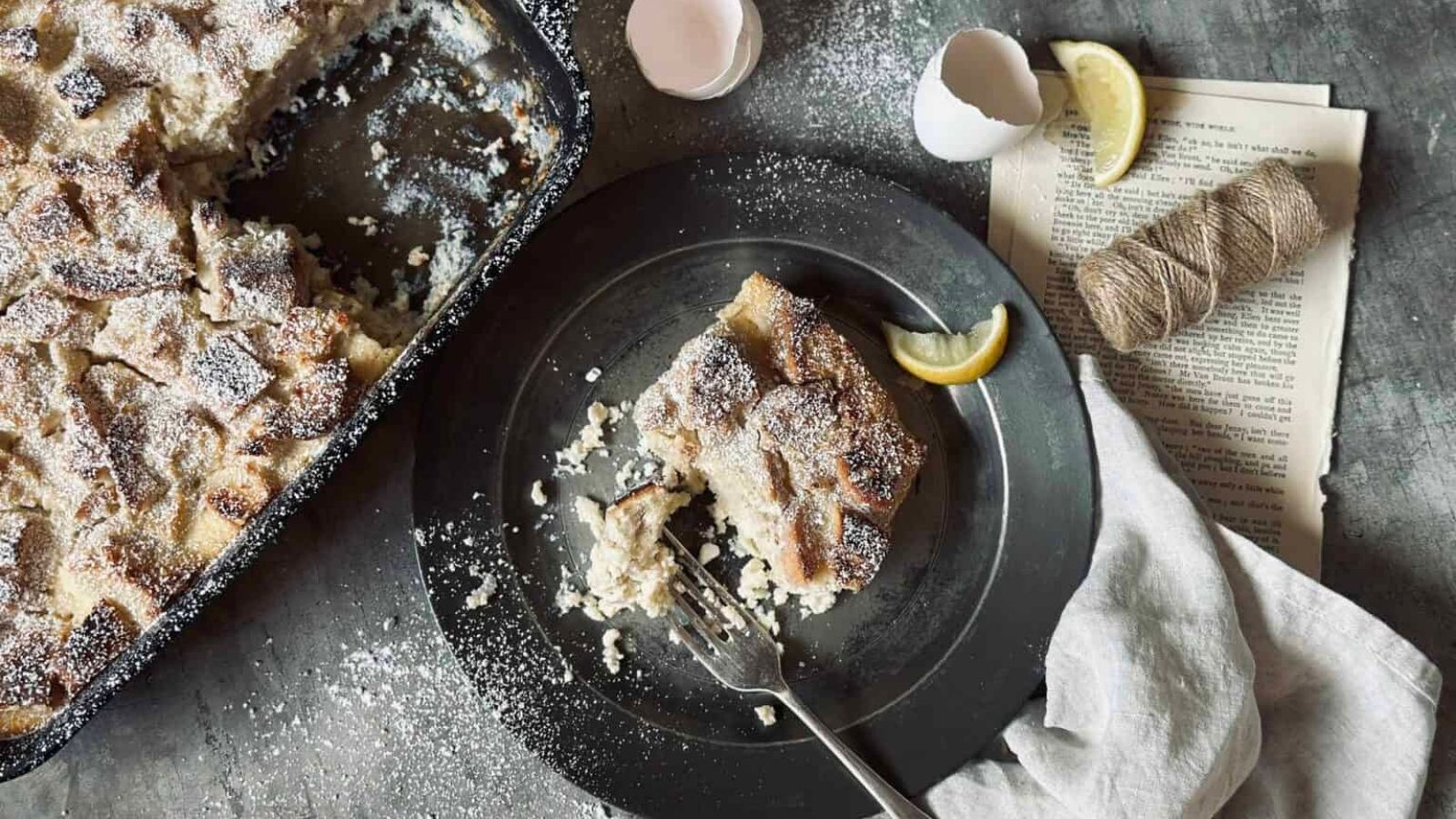 A plate with a serving of powdered sugar-dusted bread pudding, a fork, and a lemon wedge beside a baking dish, eggshells, twine, and book pages on a table.