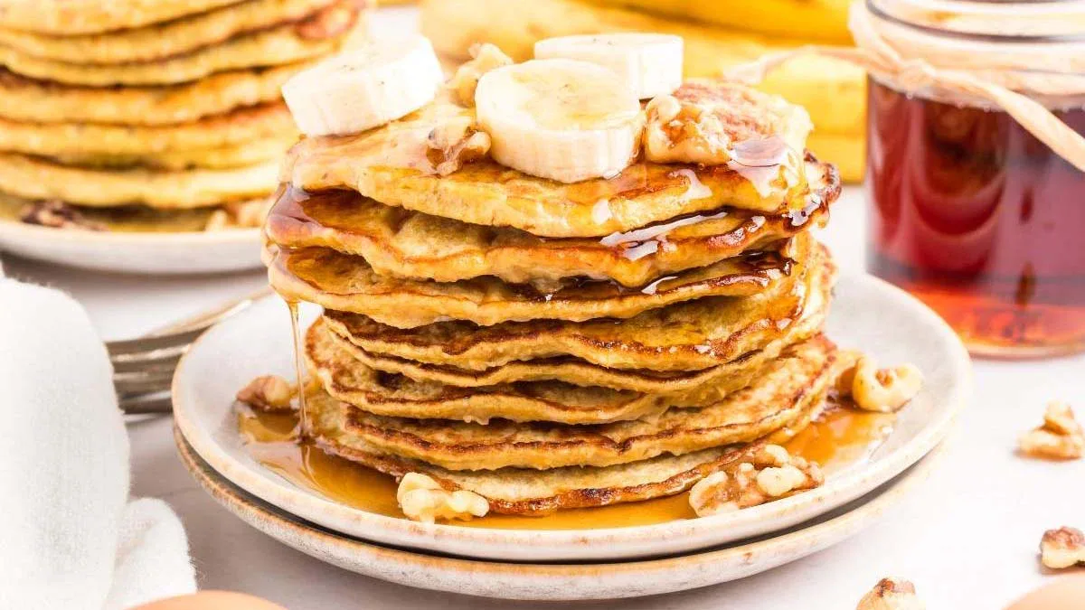 A stack of pancakes topped with banana slices and syrup, with chopped nuts scattered on the plate and a jar of syrup in the background.