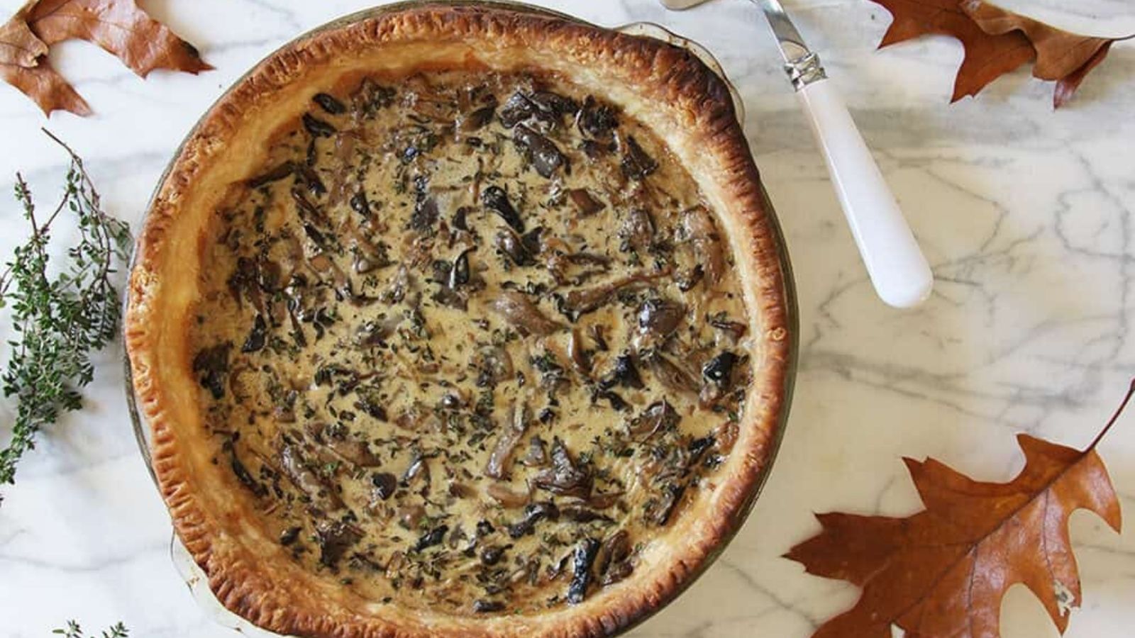 A baked savory pie with a golden crust and creamy mushroom filling sits on a marble surface, surrounded by autumn leaves and a sprig of fresh herbs.