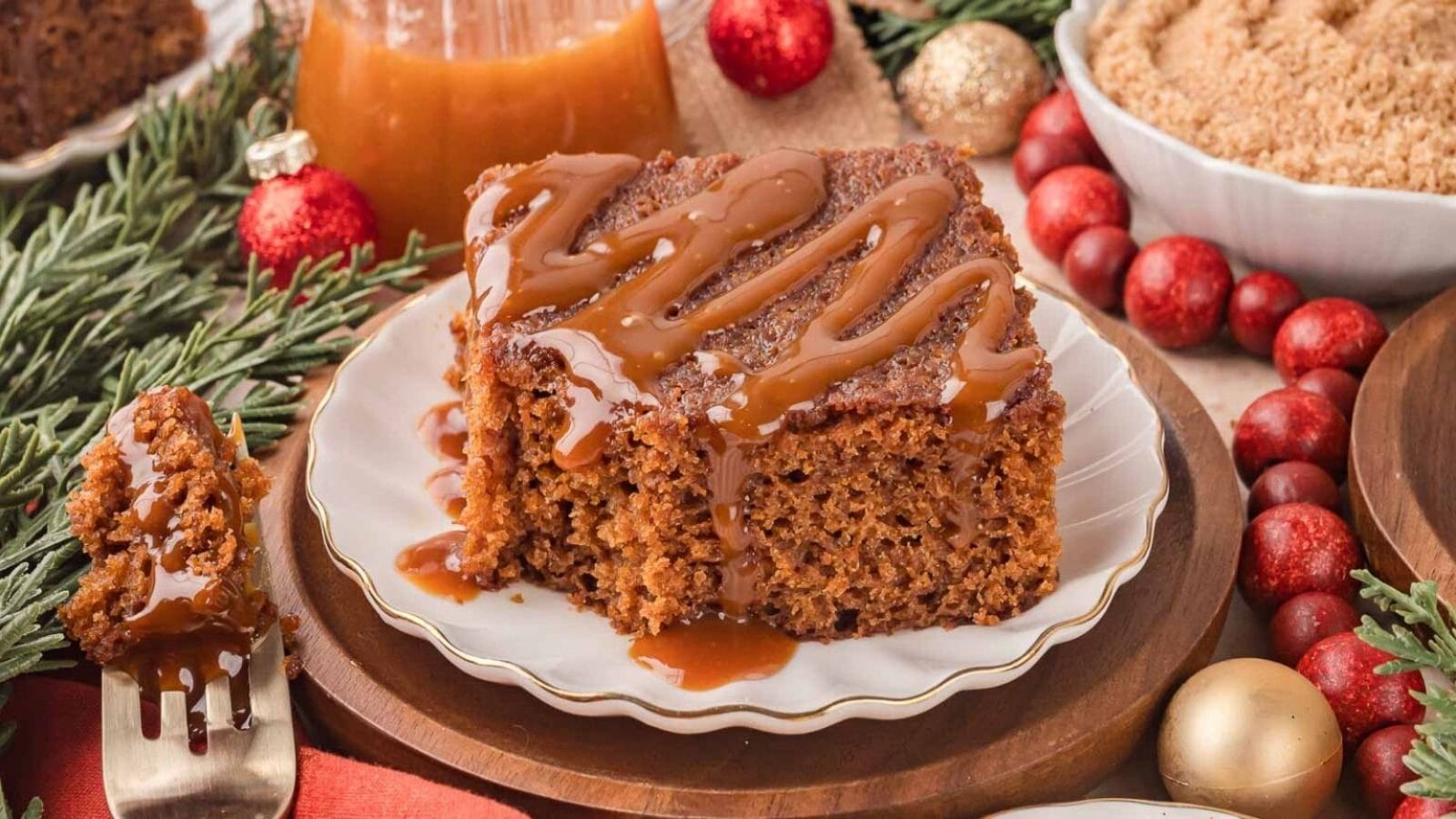 A slice of gingerbread cake drizzled with caramel sauce is served on a plate, surrounded by festive holiday decorations and ingredients.