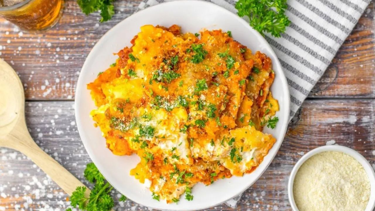 A plate of baked lasagna topped with herbs and grated cheese, placed on a wooden table with a napkin, wooden spoon, and a small bowl of grated cheese nearby.